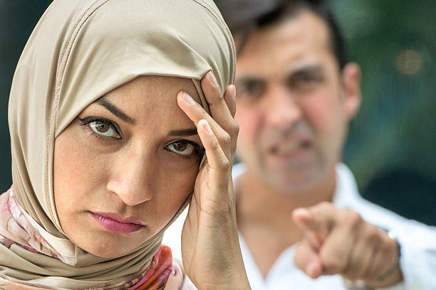 Wanita berhijab sedang memegang kepala di hadapan seorang lelaki yang sedang marah sambil menunjuk ke arah wanita di hadapannya.